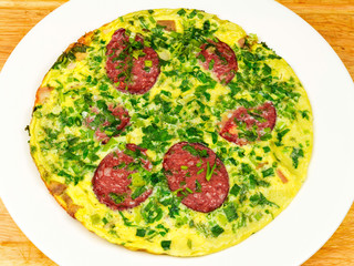 Omlete with sausage and herbs in white plate on wooden table, top view
