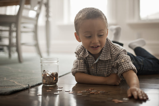 A child lying on his stomach on the floor playing with coins and putting them in a glass jar. 