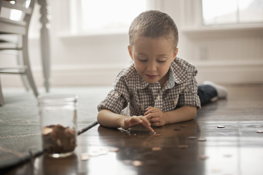 A child lying on his stomach on the floor playing with coins and putting them in a glass jar. 