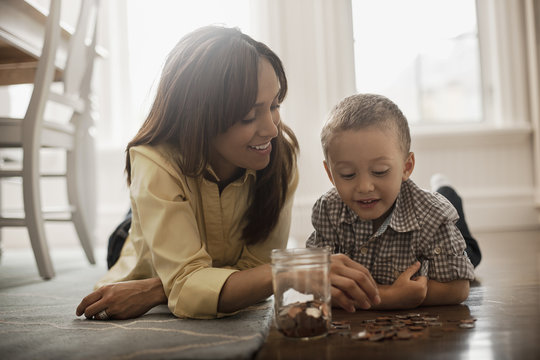 A woman and a child lying on the floor, playing with brass and silver coins, putting them into a glass jar. 