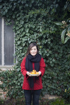 woman holding a dish full of  freshly harvested persimmons in front of a wall covered with ivy leaves