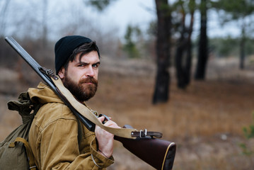 brutal hunter, bearded man in warm hat with a gun in his hand, a knife a backpack and smoking pipe in the wild forest in the autumn