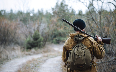 brutal hunter, bearded man in warm hat with a gun in his hand, a knife a backpack and smoking pipe...
