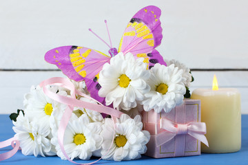 a bouquet of white daisies with a gift and a candle with butterfly on a blue wooden table