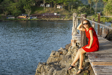 Beautiful girl in red dress sitting on the wooden bridge over the Bay in the tropics.