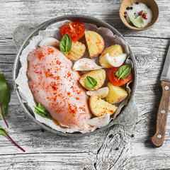 chicken breast, potatoes, onions, tomatoes - raw ingredients for making baked chicken breast with vegetables, vintage pan on a light wooden background