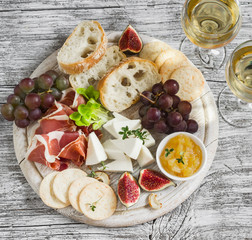 delicious appetizer to wine - ham, cheese, grapes, crackers, figs, nuts, jam, served on a light wooden board, and two glasses with white wine on bright wooden surface
