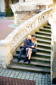 One beautiful stylish couple of young woman and senior man with long black beard sitting embracing close to each other outdoor in autumn street on stairs sunny day, horizontal picture