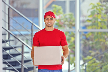 Delivery concept - postman in red uniform holding package near stairs