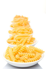 Dried pasta variety in shapes in white bowl over white background