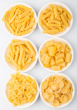 Dried pasta variety in shapes in white bowl over white background