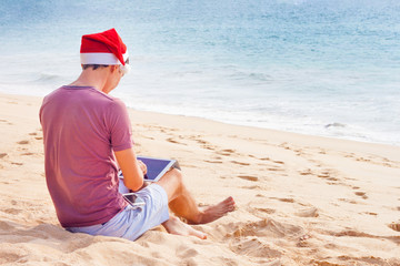 a man sitting in a Christmas hat on the sand on the beach near see and looking at the tablet, gadget, freelancer