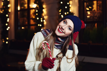 Night street portrait of smiling  beautiful young woman with Christmas candy cane. Model looking at camera. Lady wearing classic winter knitted clothes. Festive garland lights. 