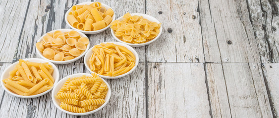Various dried pasta variety and shapes in white bowl over wooden background