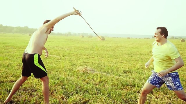 Young friends play the fool in the field during sunset with wooden rapier in slowmotion