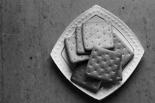 Cracker biscuits in a white ceramic square saucer on old wooden background close-up. Black and white photo. Free space for text. Copy space. Top view