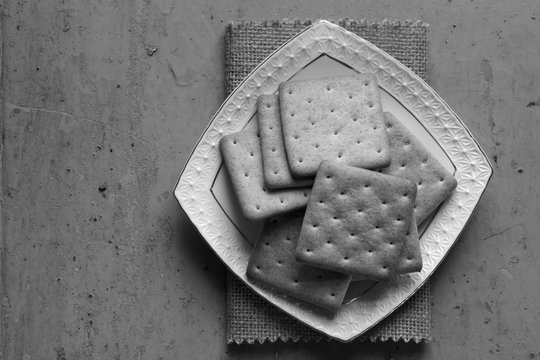 Cracker biscuits in white ceramic square saucer on a linen napkin on old painted background. Black and white photo. Free space for text. Copy space. Top view