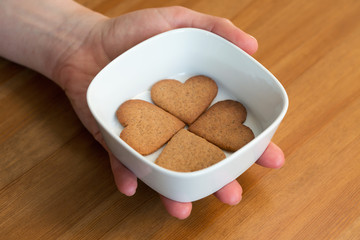A hand holding white bowl with four heart shaped homemade cookies on wooden background