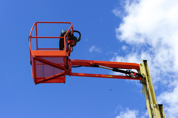 Hydraulic lift platform with bucket of construction vehicle painted in orange and beige colors with white clouds and blue sky on background 