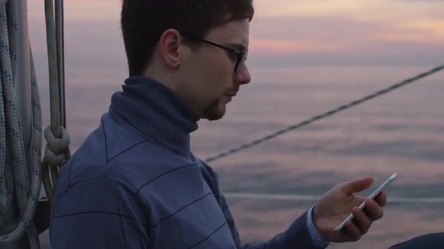 Man is looking and using phone on a sailing boat in the sea at sunset. Shot on RED Cinema Camera.