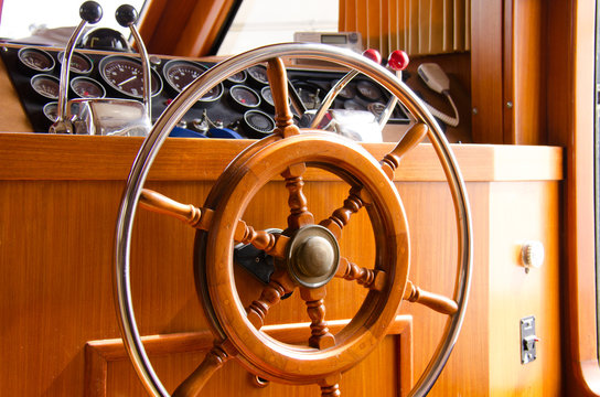 Wooden and metal steering wheel on the interior of a large yacht boat with gauges, engine throttle accelerators and vhf radio in the background