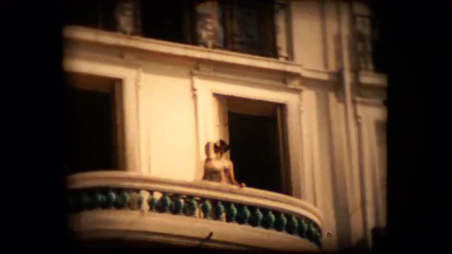 An unknown woman stands on a balcony high above a french street and waves