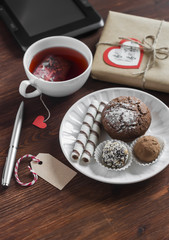 Tea cup with a homemade tea bag, sweets - cake, cookies and homemade candy, homemade Valentine's day gift in kraft paper and tablet on office wooden table.