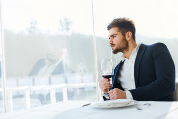 Fototapeta na wymiar Drink. Closeup Of Elegant Handsome Young Man Drinking Red Wine Sitting In Luxury Gourmet Restaurant. Businessman Relaxing And Looking Through The Window. Success Celebration Dinner Concept