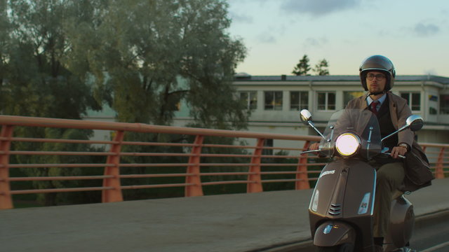 Young man in an elegant beige trench coat rides a retro scooter across a bridge at sunset