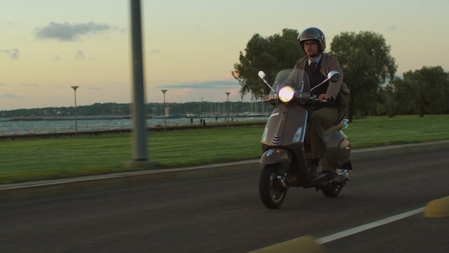 Young man in an elegant beige trench coat rides a retro scooter on a seaside road at sunset.