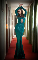 Young beautiful luxurious woman in long elegant turquoise dress posing indoors. Attractive brunette with tight fit glamorous dress in hotel lobby. Fashionable seductive female on red carpet of hall