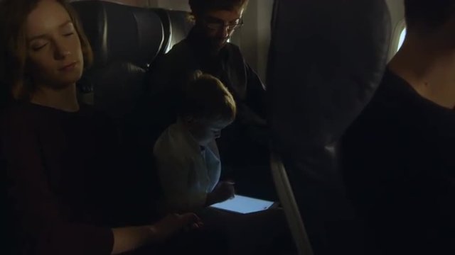 Young boy is playing with a tablet on an airplane while mother is sleeping and father sits next to a window.