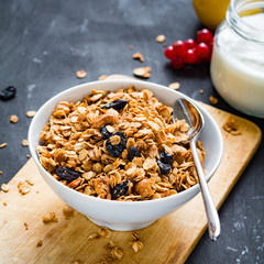 Homemade granola with nuts and dried fruits in white bowl, jar of fresh yogurt and fruits on wooden background. Square composition