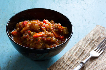 Vegetarian dish of stewed vegetables in a black ceramic bowl and fork in stainless steel on canvas on old blue table. Rustic style. Free space for text. Copy space. Selective focus