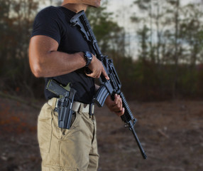 Gun owner with an assault rifle outdoors and a semi-auto pistol strapped to his side.