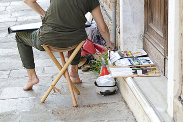 Woman artist painting on the street, she is sitting on a wooden bench seat, tools paint beside her...