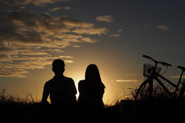 Fototapeta na wymiar A man and a woman sit beside a country road with their bicycle in the evening : silhouette photo