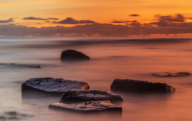 Fototapeta na wymiar Long exposure ocean sunset. Crimson sunset with waves and clouds motion blurred and flat rocks in the foreground