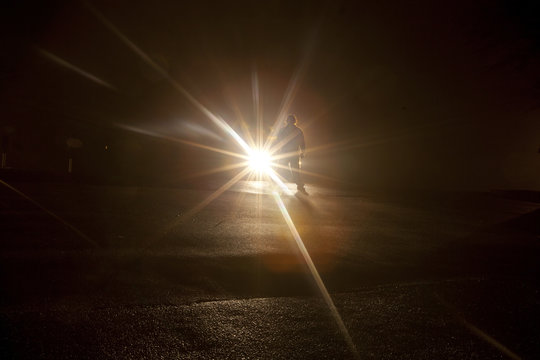 A shadowy figure standing in front of vehicle headlights in the dark of night.