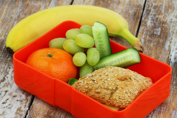 Healthy school lunch box containing whole grain roll with cheese and lettuce, cucumber sticks, grapes, mandarine and banana
