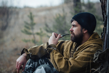brutal hunter, bearded man in warm hat with a gun in his hand, a knife a backpack and smoking pipe...