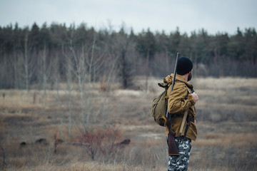 brutal hunter, bearded man in warm hat with a gun in his hand, a knife a backpack and smoking pipe...