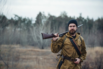 brutal hunter, bearded man in warm hat with a gun in his hand, a knife a backpack and smoking pipe...