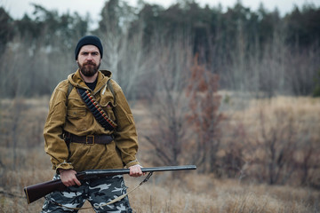 brutal hunter, bearded man in warm hat with a gun in his hand, a knife a backpack and smoking pipe...