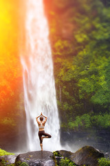 A young woman practicing yoga outdoors in front of the beautiful waterfall.  - 99105464