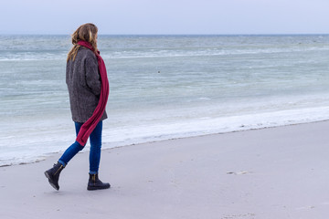 Lone sad beautiful girl walking along the shore of the frozen sea on a cold day, rubella, chicken with a red scarf on the neck