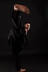 Papier Peint photo Arts martiaux Studio portrait of young karate fighter kicking over black background.