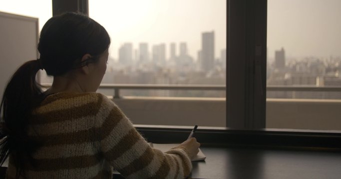 Asian women writes a journal while looking over the city
