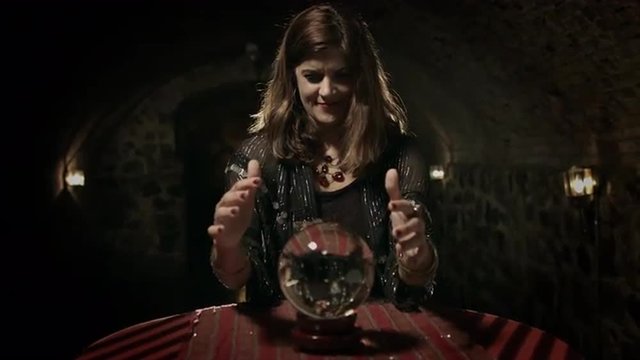 Fortune teller gazes into a crystal ball in a medieval style vaulted room.  Wide shot with jib move.  UHD 4K.