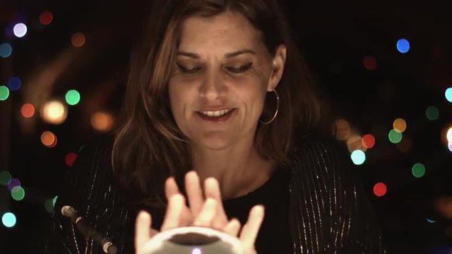 Smiling clairvoyant beckons people to join her in viewing a crystal ball.  Medium close up.